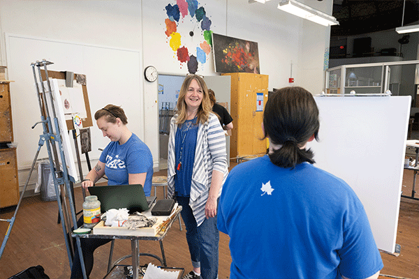 Two female students are seated in an art studio, and their female instructor stands between them. The instructor has dark blonde hair and is wearing a blue t-shirt with a grey-and-white striped, unbuttoned shirt over top of it, blue jeans, and a blue-and-white lanyard with something red hanging from the end. She is smiling at the student in the foreground, who has short back hair and whose back is turned to the camera as she sits in front of her canvas. The other student, pictured to the instructor’s left.